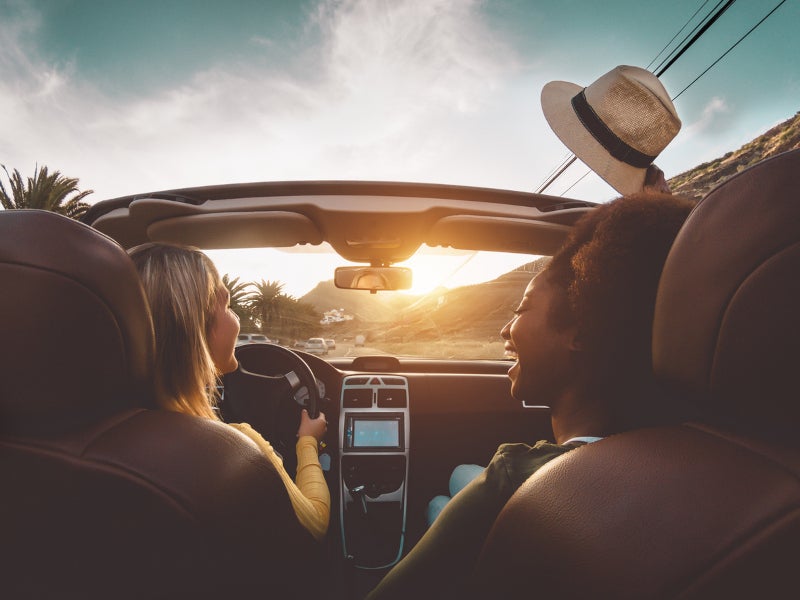 Two friends enjoying a scenic drive in a convertible car during sunset, with palm trees and hills in the background.